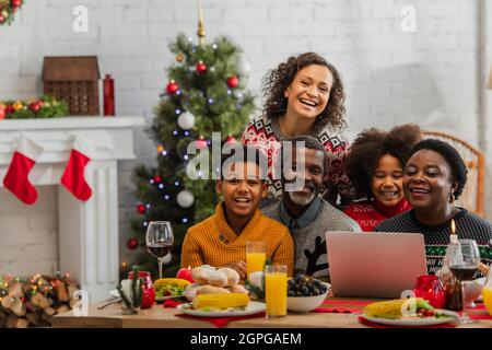 afroamerikanische Familie lächelt neben einem Laptop auf dem Tisch mit weihnachtsessen Stockfoto