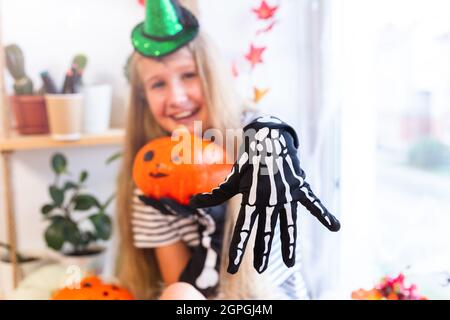 Fröhliches hübsches Mädchen 10-11 Jahre alt, verkleidet als Hexe zu Halloween, zeigt ihre Hand in einem schwarzen Handschuh mit Knochen. Trick or Treat. Selektiver Fokus. Stockfoto
