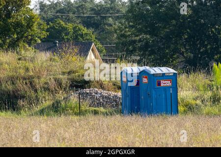 Zwei tragbare Toiletten sind in den USA blau gestrichen und befinden sich auf einem grasbedeckten Feld mit Haus im Hintergrund Stockfoto