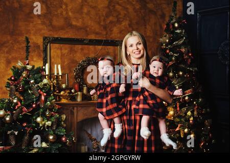 Eine glückliche Mutter mit ihren Zwillingsskindern im Neujahrsinneren des Hauses auf dem Hintergrund eines Weihnachtsbaums. Stockfoto