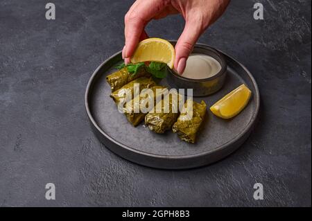 Die Hand der Frau drückt Zitrone auf Dolma mit Petersilie, Kochtopf mit saurer Creme und Zitrone auf schwarzem Teller auf dunklem Holz. Nahaufnahme. Selektiver Fokus Stockfoto