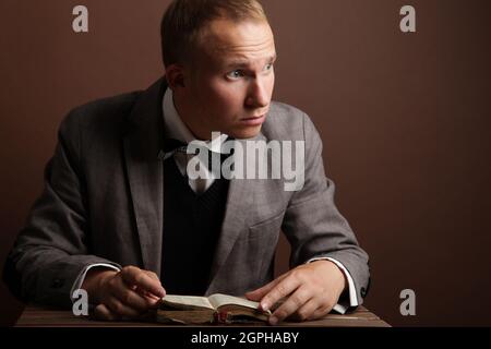 Ein gutaussehender, selbstbewusster Geschäftsmann in einem Anzug und einer runden Brille, isoliert auf einem braunen Hintergrund sitzend und ein Buch liest. Ein Mann ist nachdenkliches, überraschendes Gesicht. Hochwertige Fotos Stockfoto