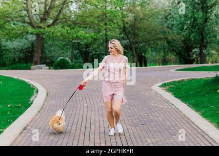 Attraktive junge Frau hält Hund spitz draußen und lächelt an der Kamera, zu Fuß im Park. Stockfoto