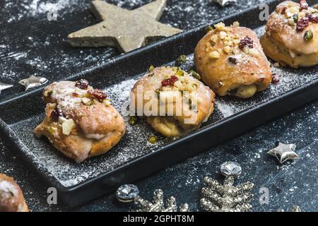 Hausgemachter Mini-Stollen mit Sahnehäubchen, getrockneten Preiselbeeren und Pistazien auf einem schwarzen Tablett auf schwarzem Hintergrund, verziert mit silbernen Sternen. Stockfoto