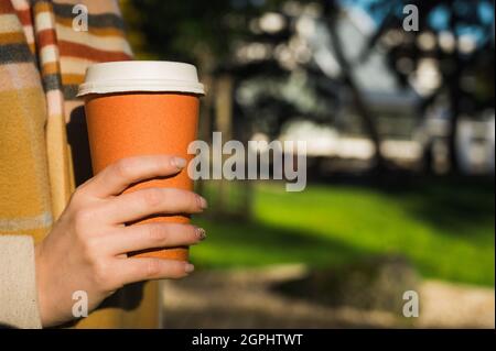 Nahaufnahme einer Tasse Kaffee aus orangefarbenem Karton in der Hand einer Frau. Getränk mitnehmen. Warmer Schal. Stockfoto