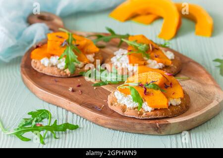 Runde Knäckebrot-Scheiben mit geröstetem Kürbis, körniger Frischkäse und Rucola auf einem Holzbrett auf hellgrünem Hintergrund Stockfoto