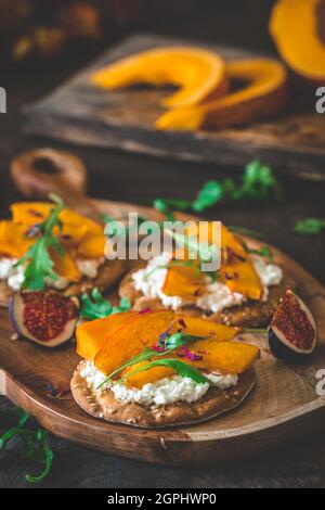 Runde Knäckebrot-Scheiben mit körnigem Frischkäse, geröstetem Kürbis und Rucola auf Holzhintergrund, vertikal Stockfoto