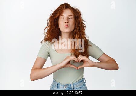 Schöne Rotschopf Frau Pucker Lippen, küssen und zeigt Herz-Geste, stehen niedlich und zart vor weißem Hintergrund Stockfoto