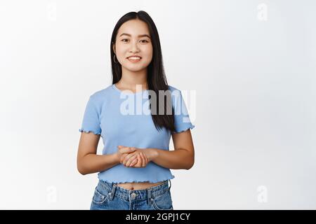 Lächelnde asiatische Frau hält die Hände zusammen und sieht freundlich aus, steht in hilfsbereiter Assistenzpose, weißer Hintergrund Stockfoto