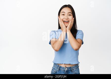 Glücklich enthusiastische asiatische Frau lächelnd, die Hände an den Wangen haltend und erfreut auf die Kamera schauend, im T-Shirt auf weißem Hintergrund stehend Stockfoto