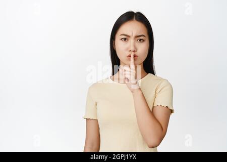 Wütend asiatische Frau shushing, zu schweigen und Stirnrunzeln, sagen, ruhig zu sein, stehend in gelbem T-Shirt über weißem Hintergrund Stockfoto