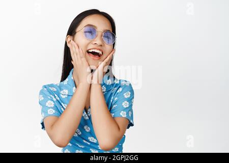 Glückliches asiatisches Mädchen, das lacht, Sonnenbrille trägt und sorglos aussieht, in Kleid über weißem Hintergrund stehend Stockfoto