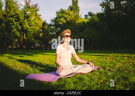 Yoga im Park. Frau mittleren Alters in Lotuspose sitzt auf Yoga-Matte auf grünem Gras. Konzept der Ruhe und Meditation. Yoga im Freien. Ältere Frau meditierende Frau Stockfoto