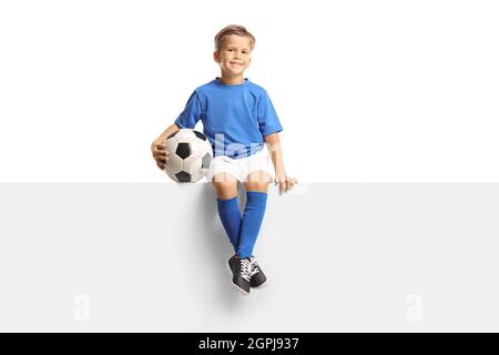 Niedlicher kleiner Junge in einem blauen Fußballtrikot sitzt auf einer leeren Platte und hält einen Ball isoliert auf weißem Hintergrund Stockfoto