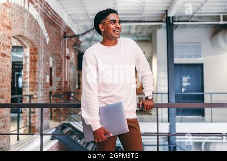 Lächelnder Geschäftsmann, der in einem modernen Büro steht. Glücklicher junger Geschäftsmann, der einen Laptop in der Hand hält und sich an Metallgeländer lehnt. Moderne Businessman-Gebühr Stockfoto