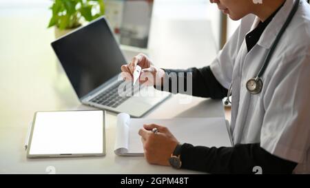 Zugeschnittenes Bild eines jungen asiatischen Arztes, der ein klinisches Thermometer in der Hand hält und Notizen auf einem medizinischen Clipboard schreibt. Stockfoto