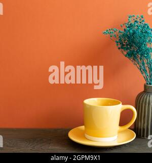 Gelbe Kaffeetasse und Vase mit grünen trockenen Blumen auf Holztisch. Roter Wandhintergrund Stockfoto