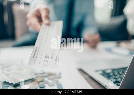 Eine Geschäftsfrau, die dem Kunden einen Scheck übergibt, und eine Rechnung, eine Münze, einen Laptop und ein Diagramm auf dem Schreibtisch im Büro. Zahlung per Scheck, Gehaltsscheck, Bezahlung Stockfoto