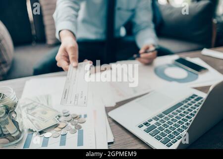 Eine Geschäftsfrau, die dem Kunden einen Scheck übergibt, und eine Rechnung, eine Münze, einen Laptop und ein Diagramm auf dem Schreibtisch im Büro. Zahlung per Scheck, Gehaltsscheck, Bezahlung Stockfoto
