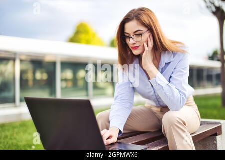 Junge Nerd Rotschopf kaukasische Frau auf Laptop lesen, im Freien im Stadtpark Stockfoto