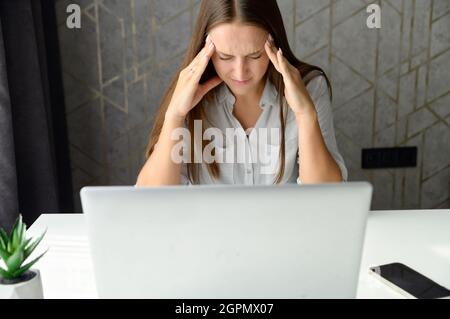 Verärgert und frustriert junge Frau mit Laptop für Remote-Arbeit von zu Hause aus, nervös über Fehler, stressigen Job, weibliche Studentin hat Problem mit Aufgabe halten Kopf vor Verzweiflung Stockfoto