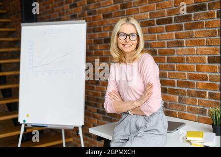 Lehrerin mittleren Alters, Trainerin in einer Brille mit blondem Haar, die auf die Kamera schaut, Online-virtuelle Lektionen per Zoom-Telefonkonferenz gibt oder Geschäftsfrau, die das Projekt auf einem Whiteboard vorstellt Stockfoto