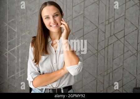 Charmante optimistische Frau, die auf dem Smartphone spricht und auf den grauen Hintergrund die Kamera anschaut, Unternehmerin steht im Heimbüro und telefoniert, chattet am Handy Stockfoto