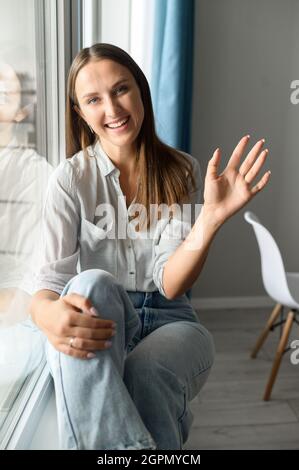 Ich freue mich auf Bis zum nächsten Mal. Freundliche, entzückende junge Frau winkt freudig der Kamera zu, lächelt und grüßt, macht ein Hello-Zeichen, grüßt die Teilnehmer des Online-Meetings, Vorderansicht, vertikaler Kopfschuss Stockfoto
