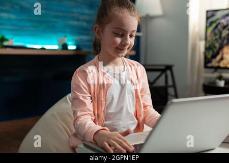 Lächelndes Schulkind, das Informationen über Laptops durchsucht, die bei Schulaufgaben arbeiten, mithilfe der elearning-Plattform für Fernunterricht. Kind sitzt auf einer großen Bohne im Wohnzimmer und studiert Literaturunterricht Stockfoto