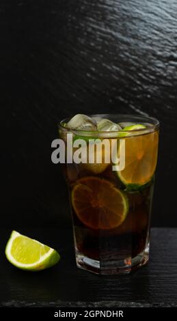 Cuba Libre Cocktail mit braunem Rum, Zitronensaft, Cola und Eiswürfeln, garniert mit einer Scheibe frischer Limette auf einem modernen Tablett aus schwarzem Stein. Bar-Konzept Stockfoto