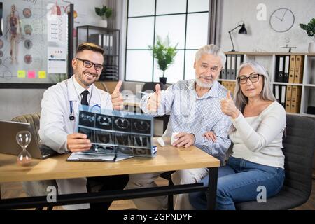 Reifes, glückliches Paar, das Daumen hoch zeigte und ihren Arzt zur regelmäßigen Untersuchung in einer modernen Klinik aufgesucht hat. Der Arzt hält einen Röntgen-CT-Scan ab, lächelt und blickt mit den Patienten auf die Kamera Stockfoto