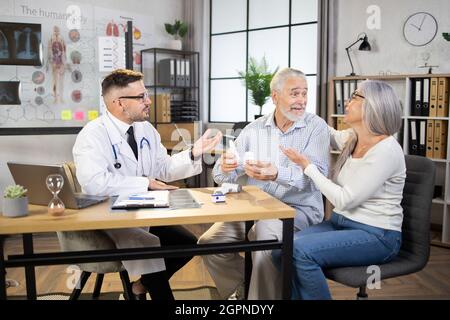 Kaukasischer Arzt in weißem Labormantel und Brille im Gespräch mit glücklichem Seniorenpaar in seinem Privatbüro. Reifer Ehemann und Ehefrau bekommen professionelle medizinische Beratung in der modernen Klinik. Stockfoto