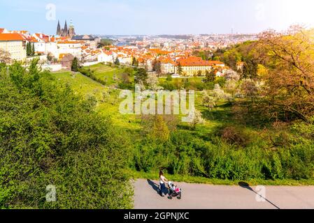 Mutter mit Kinderwagen, die im Park läuft. Blick auf die Prager Strahov-Gärten mit der Prager Burg im Hintergrund. Sonniger Frühlingstag in Prag, Tschechische Republik Stockfoto