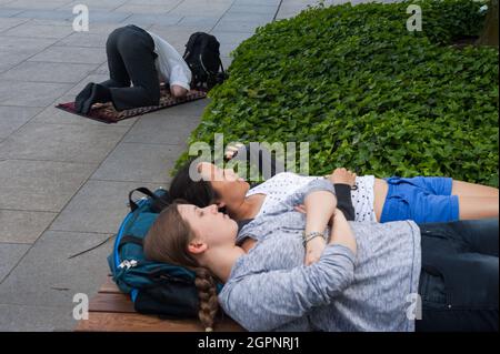 10.06.2016, Berlin, Deutschland, Europa - muslimischer Mann betet während des Ramadan auf dem Straßenbelag neben dem Universitätsgebäude, wobei Frauen auf der Bank ruhen. Stockfoto