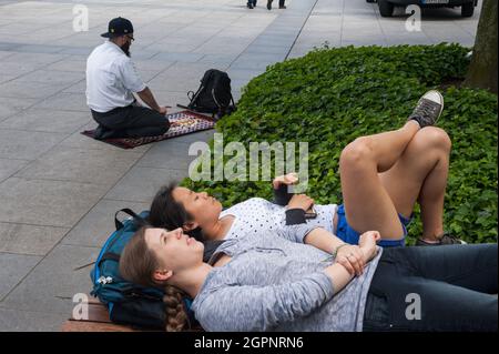 10.06.2016, Berlin, Deutschland, Europa - muslimischer Mann betet während des Ramadan auf dem Straßenbelag neben dem Universitätsgebäude, wobei Frauen auf der Bank ruhen. Stockfoto