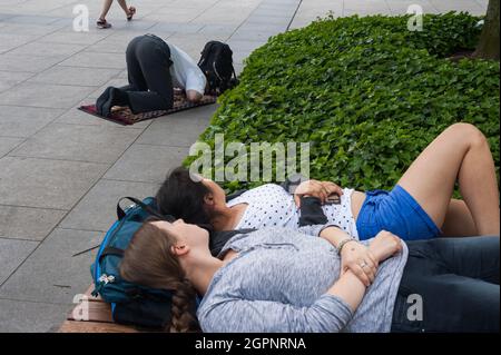 10.06.2016, Berlin, Deutschland, Europa - muslimischer Mann betet während des Ramadan auf dem Straßenbelag neben dem Universitätsgebäude, wobei Frauen auf der Bank ruhen. Stockfoto