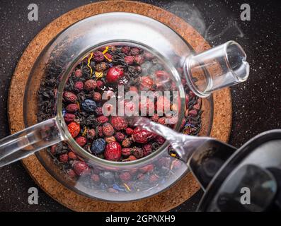 Heißes Wasser in die Glas-Teekane mit getrockneten Beeren und Blumen aus der Nähe gießen. Draufsicht. Stockfoto