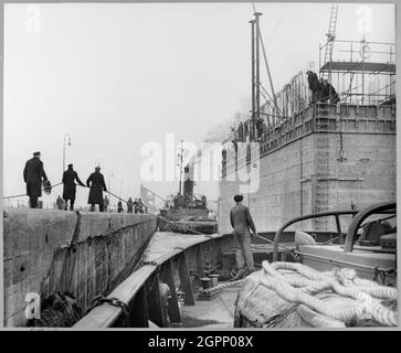 Ein Blick von einem Boot aus, der den Kühlwasserkaisson für die Coryton Ölraffinerie zeigt, der die Tilbury Docks verlässt, wobei die Leute an der Seite des Schlosses Seile zur Führung der Struktur benutzen. Während des Zweiten Weltkriegs hatte John Laing &amp; Son Ltd einige der schwimmenden Caissons für den Hafen von Mulberry gebaut, die für die Landungen der D-Day verwendet wurden. Dieser 4,200 Tonnen schwere Betonwassereinlasskaisson wurde entlang des Hafens von Mulberry gebaut und 8 Meilen die Themse hinunter vom Tilbury Dock geschleppt, um im März 1952 als Teil der Coryton Ölraffinerie installiert zu werden. Es wurde zwischen 1951 und 1952 erbaut. Stockfoto