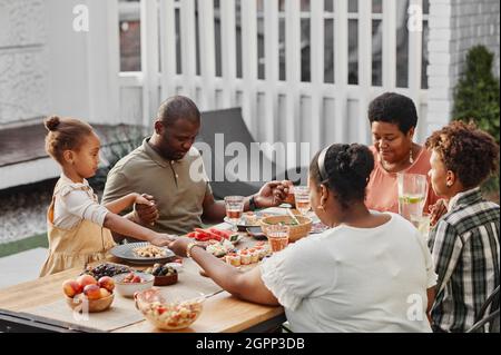 Porträt einer großen afroamerikanischen Familie, die beim Abendessen im Freien die Hände hält und betet, Platz zum Kopieren Stockfoto