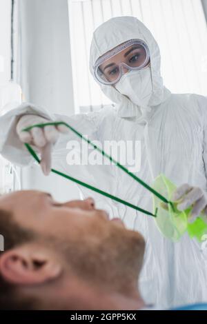 Arzt in Gefahrgut-Anzug Platzierung Sauerstoffmaske auf verschwommenen Patienten im Krankenhaus Stockfoto