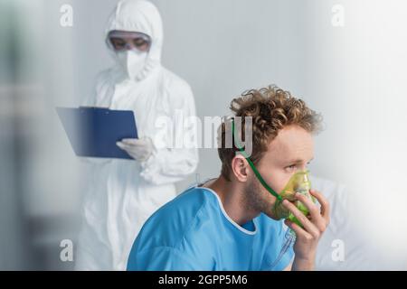 Kranker Patient atmet in Sauerstoffmaske in der Nähe verschwommen Arzt in Gefahrgut Anzug hält Zwischenablage Stockfoto