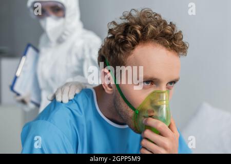 Verschwommener Arzt in Gefahrgut-Anzug zur Beruhigung des kranken Patienten beim Atmen in Sauerstoffmaske Stockfoto