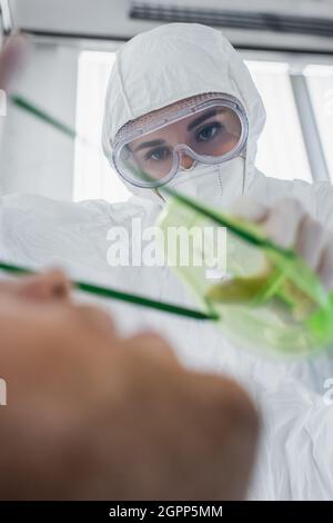 Arzt in einem Schutzanzug und einer Schutzbrille, der eine Sauerstoffmaske auf einen verschwommenen Patienten im Krankenhaus legt Stockfoto