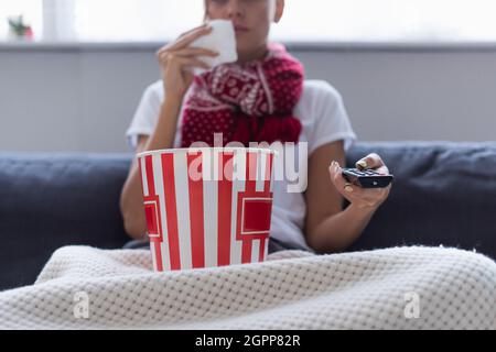 Beschnittene Ansicht einer kranken Frau mit Fernbedienung und einem Eimer Popcorn, der vor verschwommenem Hintergrund fernsieht Stockfoto