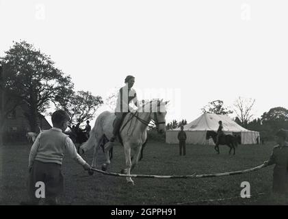 In den 1950er Jahren, historisch, draußen auf einem Feld bei einem Reitsport-Event, eine Reiterin auf einem Pferd, die gerade dabei ist, über einen hölzernen Stock zu springen, der von zwei Jungen, West Sussex, England, Großbritannien, gehalten wird. Stockfoto