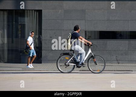Ein junger Mann geht die Straße entlang und hört Musik mit seinen Kopfhörern, während ein anderer Mann an einem Sommertag auf einer Straße Fahrrad fährt. Stockfoto