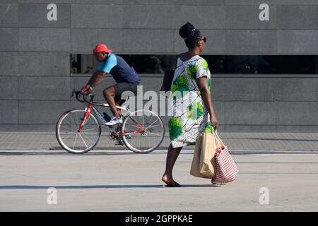 Schwarze Frau läuft, während ein schwarzer Mann im Sommer in einer Straße einer Stadt Fahrrad fährt. Stockfoto