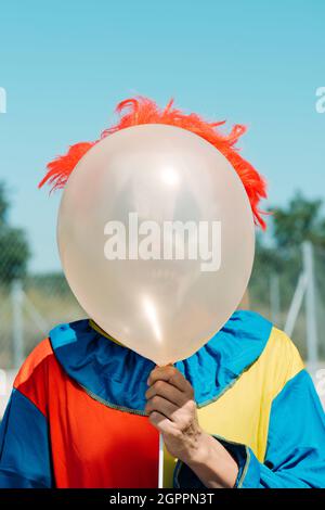 Nahaufnahme eines gruseligen Clowns, der im Freien ein farbenfrohes gelb, rot und blau gekleidtes Kostüm trägt und einen Ballon vor sein Gesicht hält Stockfoto