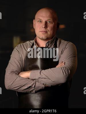 Selbstbewusster Mann in der Lederschürze faltete seine Arme, die auf dunklem Hintergrund standen und die Kamera betraten. Portrait des Handwerkers Stockfoto