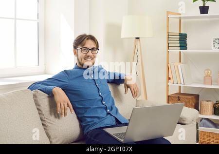 Porträt eines selbstbewussten, glücklichen jungen Mannes, der mit einem Laptop zu Hause auf dem Sofa sitzt. Stockfoto